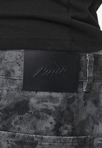 Temno sivi denim hlače z črno usnjeno oblogo na pasu, ki nosi embosirano logo. Tkanina ima marmorirano teksturo in subtilen vzorec.