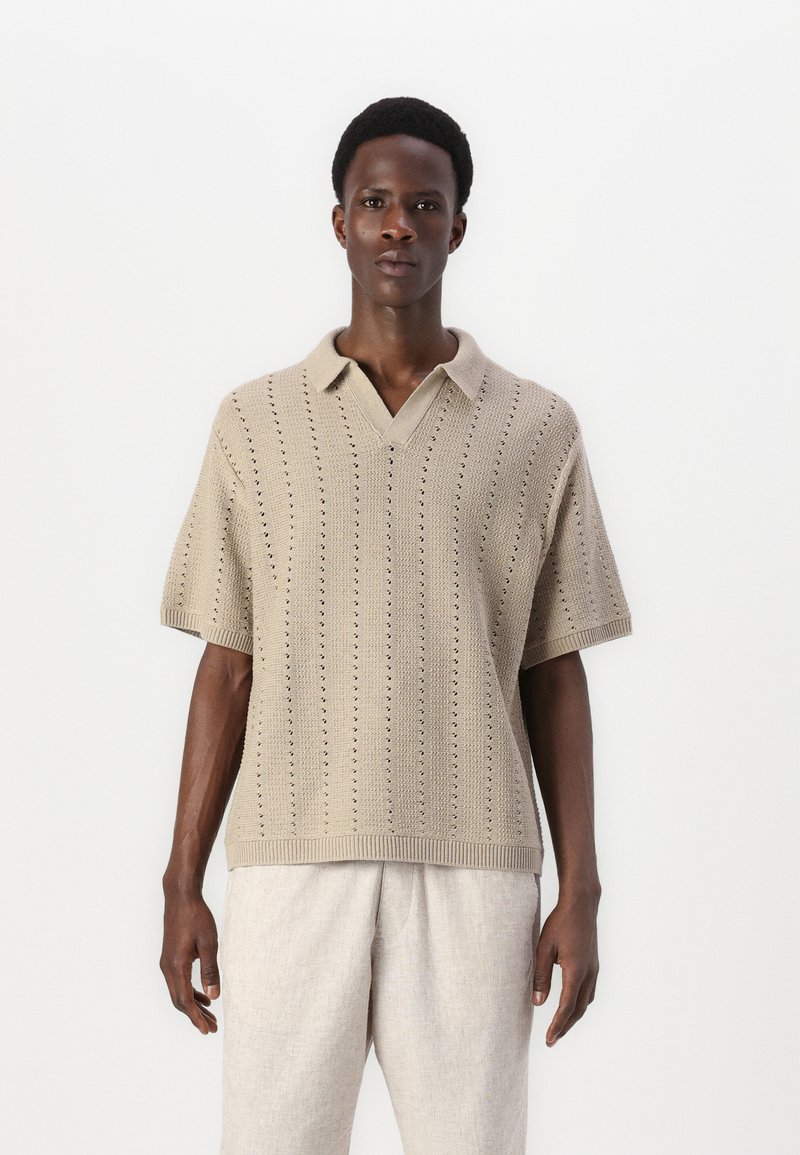Polo shirt en tricot beige avec un col en V, manches courtes et un design perforé. Porté avec un pantalon léger à coupe décontractée.