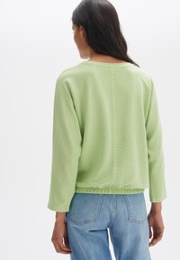 Hellgrünes, geripptes Crop-Top mit langen Ärmeln und gerafftem Saum. Die Rückseite hat eine gerade Naht. Kombination mit blauen Jeans.