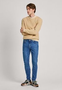 Sudadera beige, jeans ajustados azules y zapatillas multicolores. Texturas suaves con un ligero desgaste en los jeans y un ribete blanco en el cuello que contrasta.