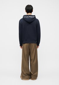 Person med kort svart hår iført en marineblå hettegenser med teksten "CLOSED" på hetten og løse, brede brune bukser, stående med ryggen til.