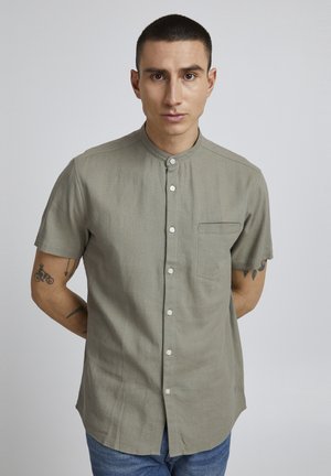 Camisa de manga corta en tejido color verde olivo claro con cuello tipo banda, cierre delantero de botones, un bolsillo en el pecho y una textura sutil.