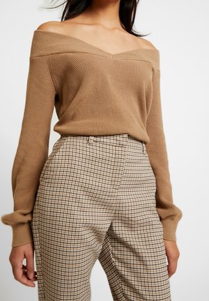 Femme portant un pull en maille camel à épaules dénudées, associé à un pantalon taille haute beige et marron à carreaux.