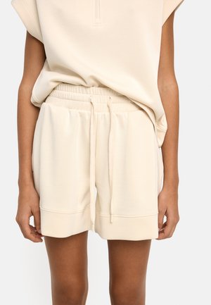 Beżowe szorty z elastycznym pasem i sznurkiem, noszone z dopasowanym luźnym beżowym topem, odsłaniające środkową część ud i przedramiona.