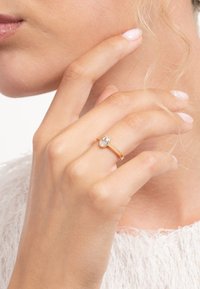 Gold ring with an oval clear stone, smooth band, worn on a hand gently touching the face. Skin tone is light, with natural nail polish on fingers.