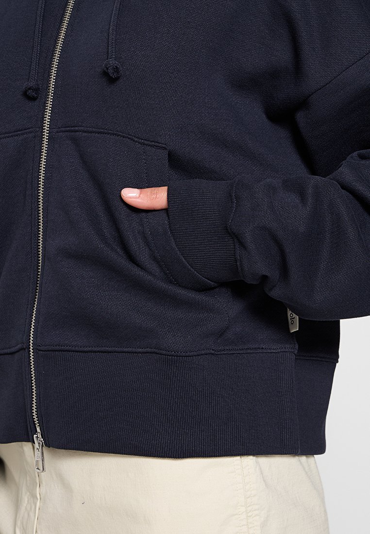 Ręka w kieszeni granatowej bluzy z zamkiem, z prążkowanymi ściągaczami i sznurkiem, noszonej na beżowych spodniach.