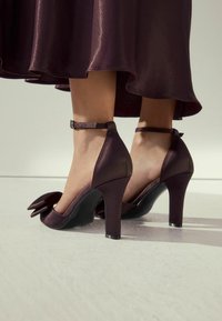 Donkerpaarse high heels met een strikdetail, gemaakt van gladde satijn, met een enkelbandje en een chunky hak.