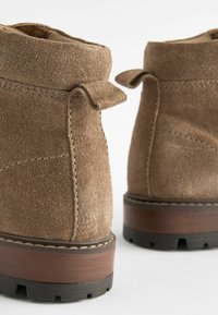 Bottines en daim marron avec une surface texturée, présentant un bout arrondi et un petit talon robuste. Comprend des tirants décoratifs à l'arrière.