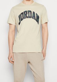 Beżowa koszulka z bawełny z dużą, zakrzywioną grafiką "JORDAN" w ciemną kratę. Krótkie rękawy i okrągły dekolt. Gładka tekstura.