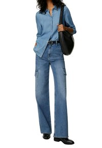 Lichtblauwe denimoverhemd gecombineerd met wijde blauwe jeans met cargozakken, een zwarte riem en een zwarte schoudertas. Zwarte enkelboots.