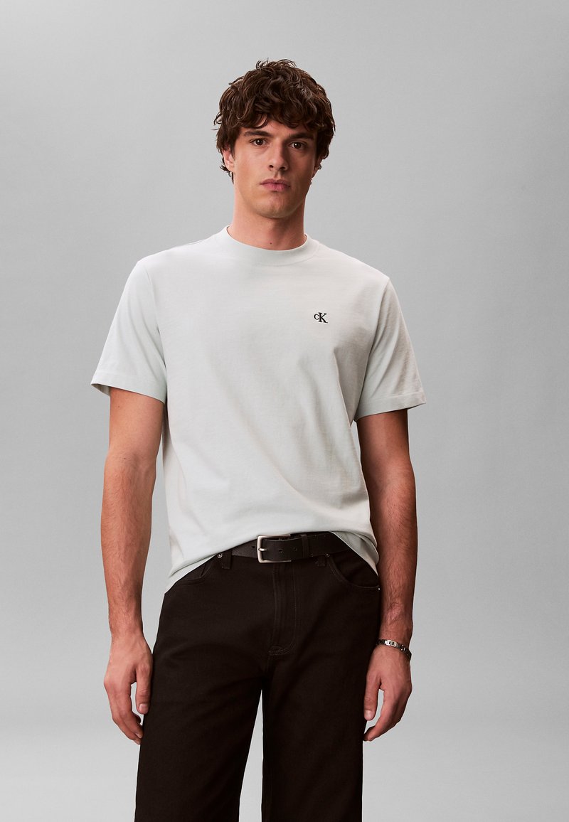 Jeune homme aux cheveux bouclés portant un t-shirt Calvin Klein gris clair et un pantalon noir sur un fond gris uni.