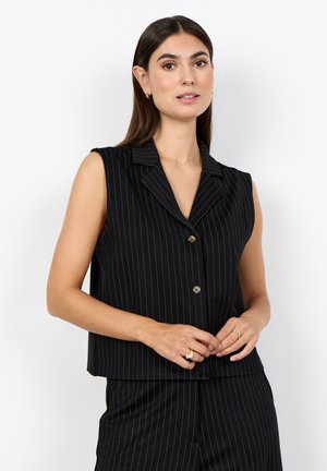 Woman with long brown hair wears black sleeveless pinstripe blazer and matching trousers, standing against a plain white background.