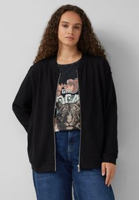 Veste zippée noire en tissu texturé, avec col rond et poignets côtelés, superposée à un t-shirt graphique avec un motif de tigre, associée à un jean bleu.