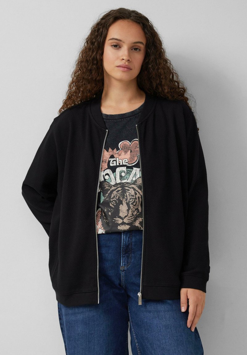 Zwarte ritsjack met structuurstof, ronde kraag en geribbelde manchetten, laag over een graphic t-shirt met een tijgerprint, gecombineerd met blauwe jeans.