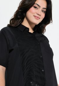 Zwarte shirt met korte mouwen, voorzien van een gestructureerd transparant zwart paneel met een zebrapatroon. Knopsluiting aan de voorkant en een enkele borstzak.