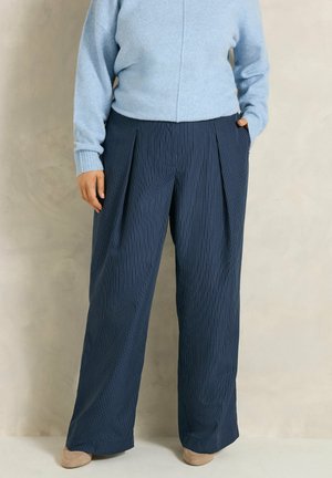 Personne porte un pull bleu clair et un pantalon large bleu foncé à rayures fines, debout devant un fond neutre.
