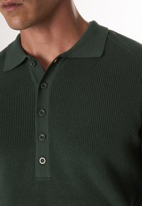 Dunkelgrünes gestricktes Polohemd mit ribbeliger Textur, spitzem Kragen und einem Knopfverschluss mit sechs schwarzen Knöpfen.