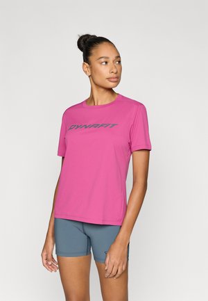 Rosa sportliches Kurzarmshirt mit "DYNAFIT"-Logo in Dunkelgrau, aus leichtem Stoff hergestellt. Kombiniert mit blauen Sportshorts.