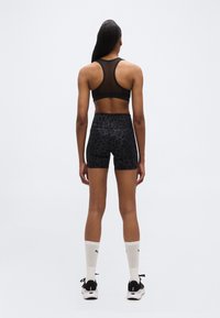 Γυναίκα που στέκεται με την πλάτη, φοράει μαύρο αθλητικό σουτιέν από δίχτυ, μαύρο σορτς με λεοπάρ σχέδιο, λευκές κάλτσες και μαύρα αθλητικά παπούτσια σε λευκό φόντο.