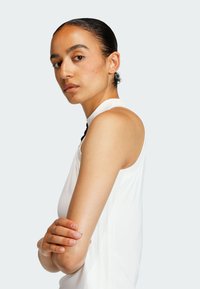 Biely top bez rukávov s vysokým výstrihom, s hladkou textúrou a jemnými vertikálnymi švovými detailmi. Model má prekrížené ruky.