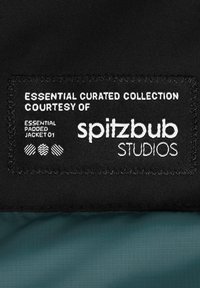 Zwarte stoffen label met witte tekst "ESSENTIAL CURATED COLLECTION", bevat "spitzbub STUDIOS" en productdetails op een groene achtergrond.