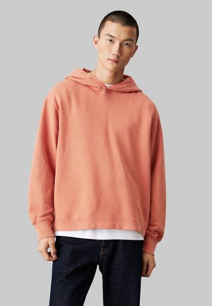 Jeune homme portant un sweat à capuche corail ample par-dessus une chemise blanche et un jean foncé, sur un fond gris clair uni.