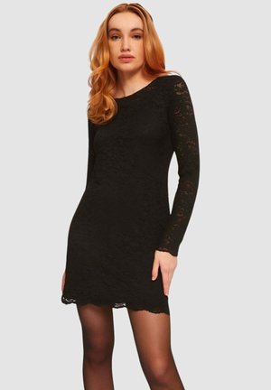 Donna con capelli rossi che indossa un vestito nero aderente di pizzo a maniche lunghe e calze nere trasparenti su uno sfondo semplice.