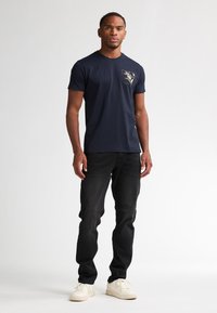 Navyblauwe katoenen t-shirt met een bloemenafbeelding op de linkerkant van de borst, gecombineerd met zwarte jeans en witte sneakers. Eenvoudig en casual ontwerp.