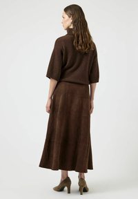 Top in maglia marrone con colletto alto, abbinato a una lunga gonna fluida in suede marrone. Il modello indossa scarpe con tacco alto color taupe. Texture liscia.