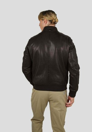 Homme aux cheveux blonds attachés en chignon, portant une veste en cuir marron foncé et un pantalon beige, vu de dos sur un fond gris uni.