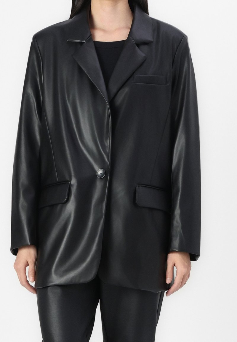 Femme portant un blazer en cuir noir brillant avec un bouton, des poches à rabat, et un pantalon noir assorti.
