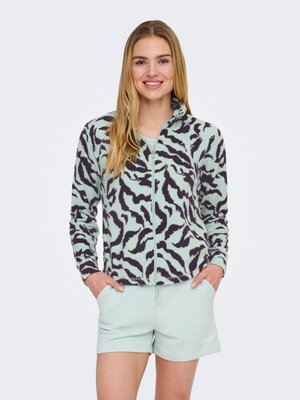 Donna sorridente con lunghi capelli biondi che indossa una giacca con zip verde menta e nera con motivi e pantaloncini verde chiaro, con le mani nelle tasche.