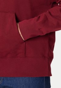 Felpa con cappuccio color bordeaux con tasca frontale, polsini a costine e una texture morbida. Ambientata su sfondo di denim scuro, che ne mette in risalto la ricchezza del tessuto.