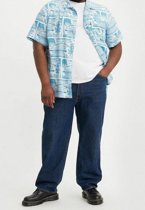 Homme portant un jean bleu foncé, des chaussures noires, un t-shirt blanc et une chemise à manches courtes avec un texte bleu et un imprimé graphique.