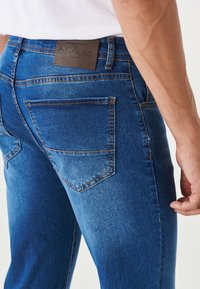 Jeans in denim blu scuro con una vestibilità tailored, dotati di tasche posteriori con cuciture visibili e un'etichetta in pelle marrone in vita.