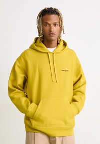 Sweatshirt à capuche jaune avec une poche frontale et un cordon de serrage, arborant un petit logo brodé. Texture douce en coupe décontractée.