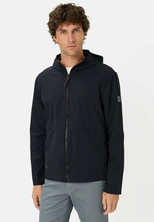 Man met krullend haar, gekleed in een zwarte capuchonjas met rits en grijze broek, staand tegen een effen lichte achtergrond.