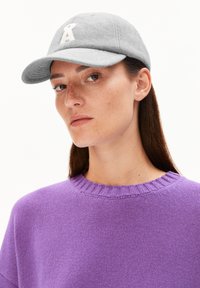 Femme avec de longs cheveux bruns portant une casquette grise avec un logo blanc et un pull en laine violet, légèrement tournée vers la gauche sur un fond blanc.
