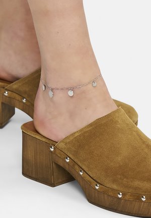 Silberne Kettenankelkette mit kleinen ovalen Anhängern, am Knöchel getragen. Sie verfügt über einen hellbraunen Wildleder-Clog mit Holzabsatz und silbernen Nietenakzenten.