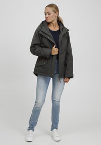 Donkergrijze hooded jack met een gestructureerde afwerking en houten accentknopen. Draagt met lichtblauwe spijkerbroek en witte sneakers.