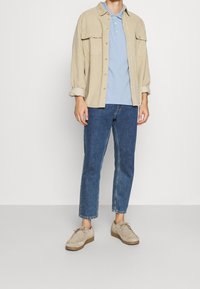 Camicia a coste color beige chiaro con due tasche, polo blu chiaro, jeans blu scuro e scarpe beige con suola marrone. Outfit casual a strati.