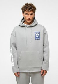 Grijze oversized hoodie met een voorzak, trekkoord in de capuchon, blauwe logo patch en witte tekst "Essentials" op de linker mouw. Soepele textuur.