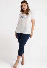 Camiseta blanca de manga corta y escote en V con "La Dolce Vita" impreso en color marino, combinada con leggings recortados en color marino y sandalias plateadas con tiras.