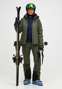 Skioutfit mit einer olivgrünen Jacke und Hose, blauen Skischuhen, schwarzem Helm und reflektierenden Skibrillen, die schwarze Skier mit Bindungen hält.