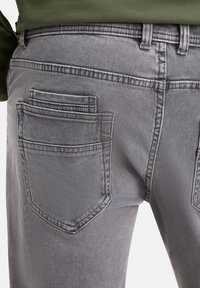 Jeans en denim gris avec une finition texturée, dotés d'une poche arrière unique et de détails de couture. La ceinture comporte des passants pour une ceinture.