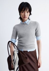 Kurzärmliger grauer Kaschmirpullover über einem hellblauen Hemd mit Kragen, kombiniert mit einer dunkelbraunen Handtasche, die mit einem Seidenschal verziert ist.