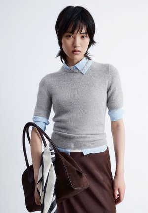 Short-sleeve grey cashmere sweater over a light blue collared shirt, paired with a dark brown handbag featuring a silk scarf.