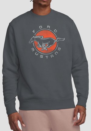 Man wearing dark gray sweatshirt with a vintage-style orange circle and running horse logo with "Ford Mustang" text.