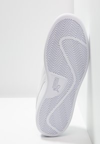 Suola di una sneaker Puma bianca che mostra un motivo a zigzag testurizzato e il logo Puma, posizionata verticalmente contro un muro bianco.