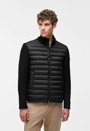 Jeune homme aux cheveux bouclés portant une veste matelassée noire zippée avec des manches longues en tricot et un pantalon beige, regardant légèrement sur le côté.